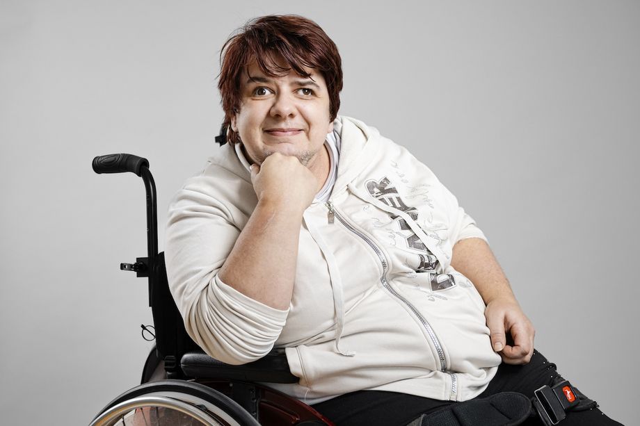 Melanie Lux hat kurze, dunkle Haare und sitzt im Rollstuhl. Sie hat ihr Kinn auf die Hand gestützt und schaut freundlich in die Kamera.