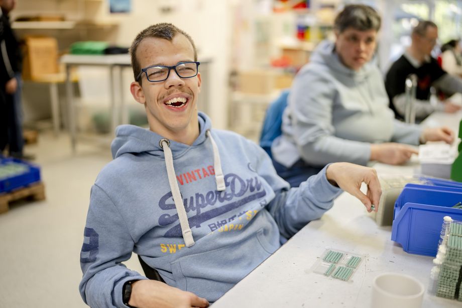 Ein junger Mann mit breitem Lächeln bei der Arbeit. Er verpackt Düngestäbchen. Neben ihm sind verschwommen andere Beschäftigte zu sehen.