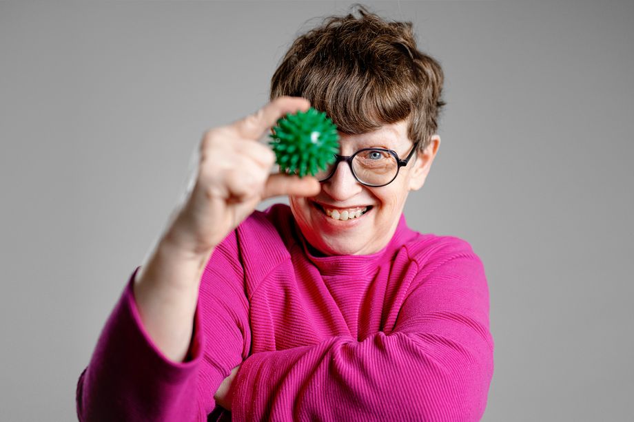 Eine Mitarbeitende von Westfalenfleiß schaut fröhlich in die Kamera und hält einen kleinen grünen Ball vor ihr rechtes Auge