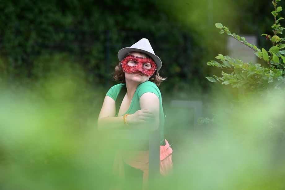 Swaantje mit einem Hut und einer Maske verkleidet, fotografiert durch einen grünen Strauch
